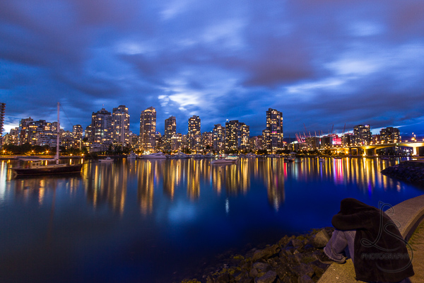 A person crouched on the waterfront as evening descends on the illuminated city of Vancouver | LotsaSmiles Photography